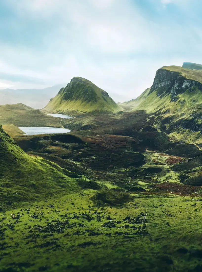 A lush, green valley with small lakes and rocky hills under a partly cloudy sky.